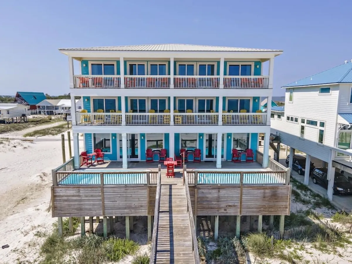 A three-story beach house featuring spacious balconies, red chairs, and blue accents. The balconies are styled for relaxation, with clear sightlines to the ocean and the pool area visible below, creating a sense of seamless indoor-outdoor living.