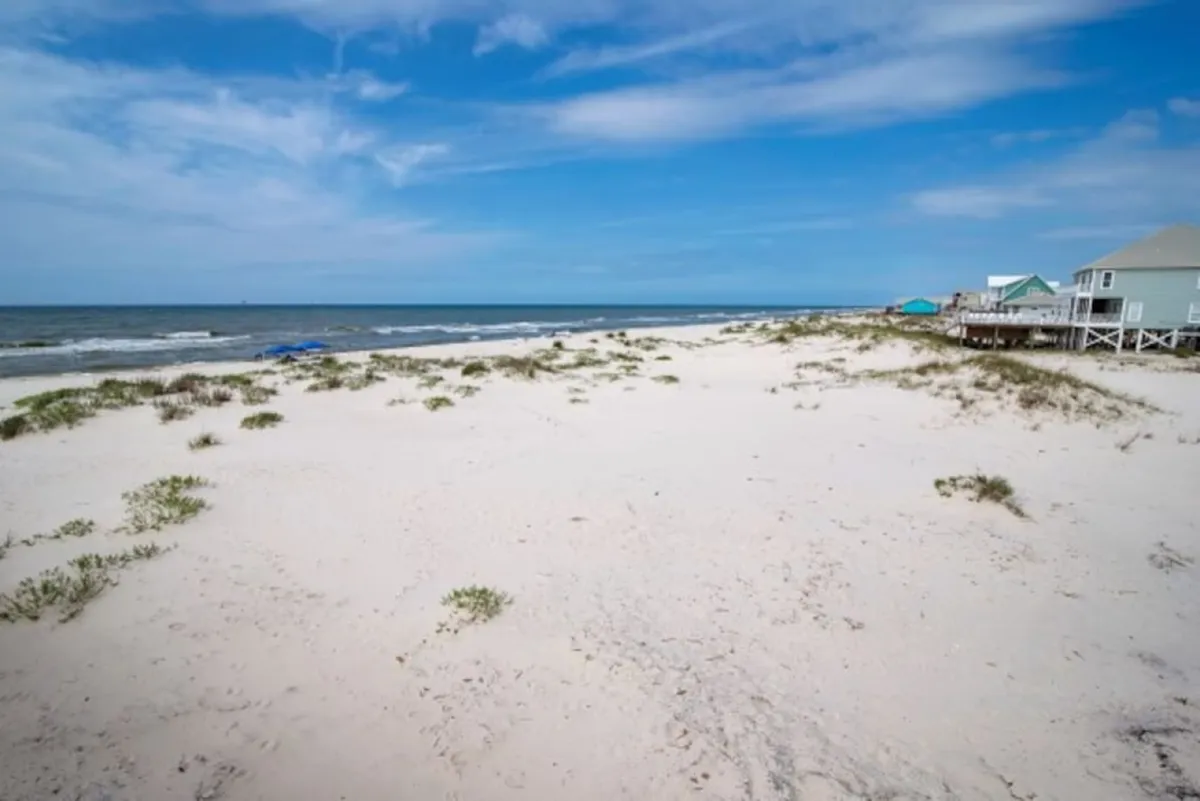 A sweeping view of a quiet sandy beach bordered by calm ocean waves, with a few distant beach houses peeking above the dunes. The shore is largely empty, evoking a sense of tranquility and space for leisurely walks or family outings.