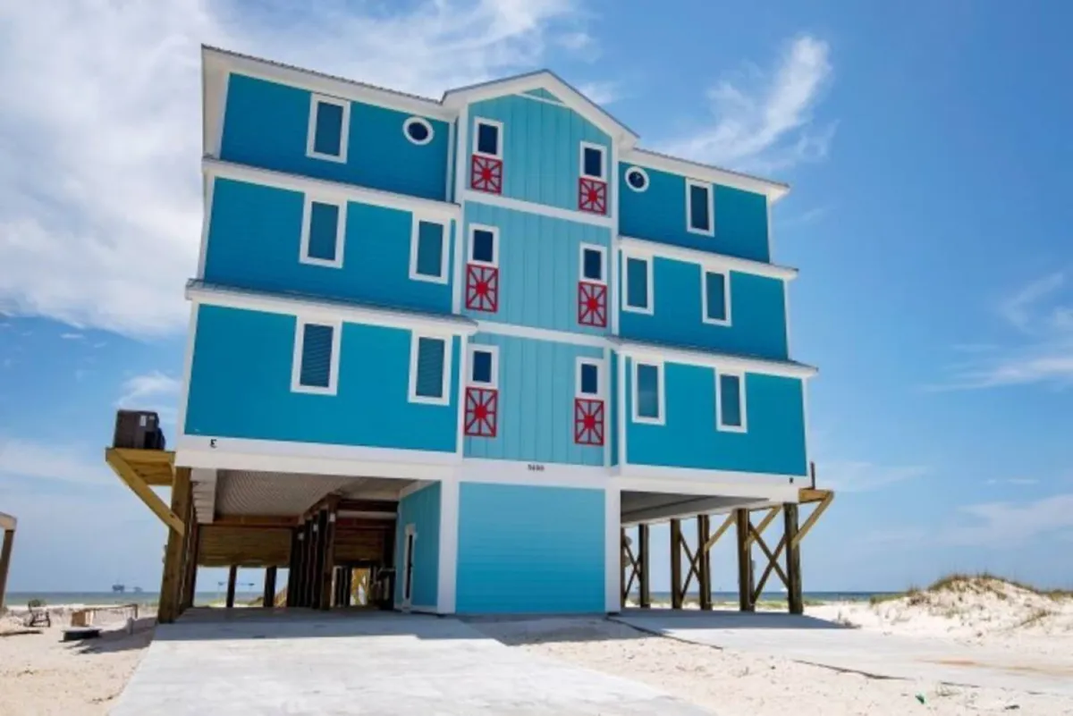 The image showcases a cheerful, bright blue beachfront house with eye-catching red shutters, perched on a stilted foundation. The inviting porch offers space to relax while taking in the Gulf views. The house sits just steps from the sandy shoreline, radiating a vibrant, welcoming vibe ideal for group gatherings and beach adventures.