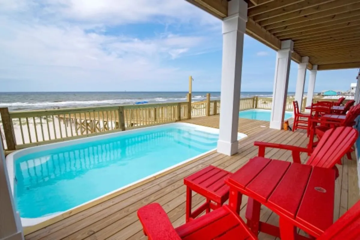 A sparkling private swimming pool surrounded by a sunny deck with bright red chairs, bordered by an ocean view in the background. The setting is inviting for swimming, sunbathing, and outdoor gatherings.