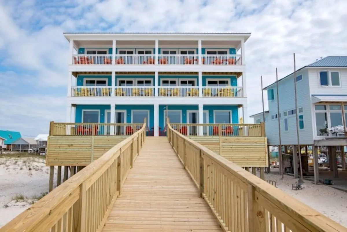 This image captures the beachfront property with colorful chairs on balconies and a wooden walkway leading down to the sand. The view emphasizes the spaciousness of the home, its proximity to the Gulf, and inviting outdoor spaces perfect for group activities and socializing.