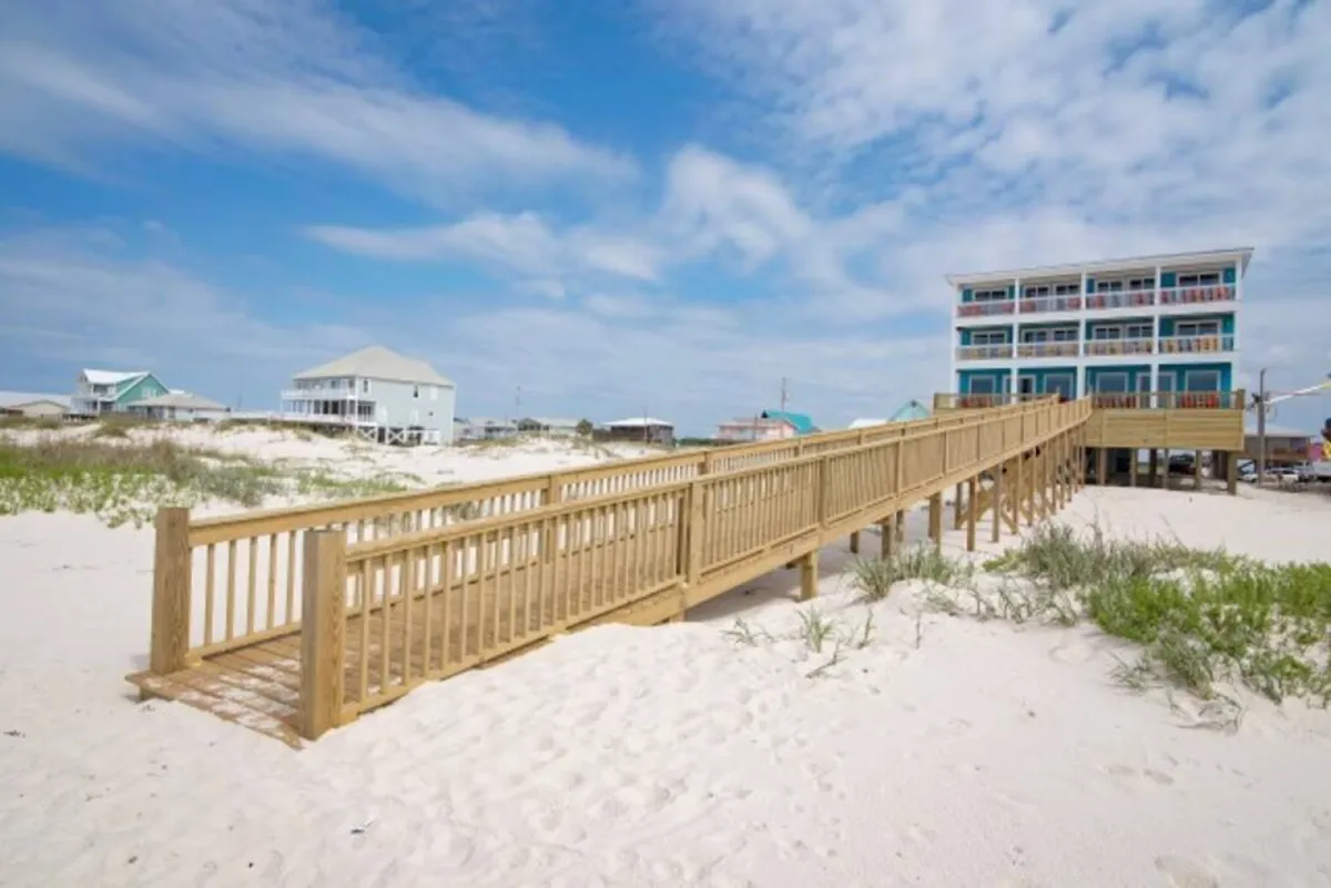 A wide exterior shot featuring a wooden walkway leading directly to a pristine beachfront property, framed by an expansive sky. The scene captures the deck space, the sandy shore, and inviting sunset lighting, emphasizing the proximity to the Gulf waters and the sense of openness perfect for celebrations.