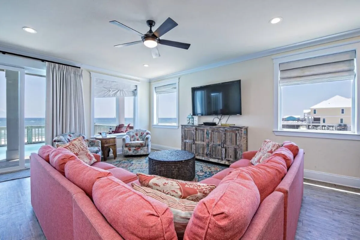A spacious and well-lit living room with a large pink sectional sofa, a round coffee table, expansive windows showcasing a sea view, and a comfortable TV unit. The space is designed for communal gatherings, relaxation, and enjoying the ocean scenery, perfect for hosting family celebrations or group events.