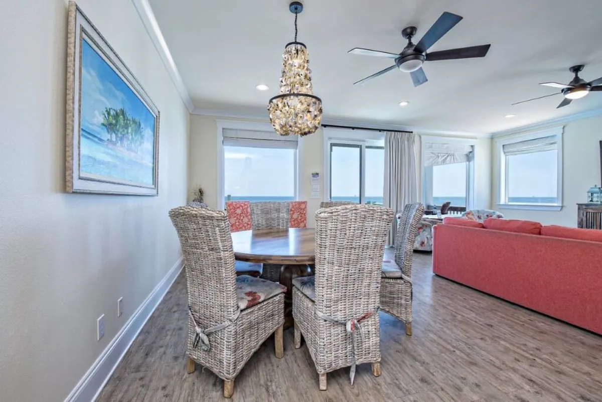 A bright dining area with a large round table surrounded by woven dining chairs, set under a stylish chandelier. Large windows reveal ocean views, and a red sofa adds a pop of color, blending relaxation and dining in one open space. The layout encourages group meals, festive celebrations, and game nights overlooking the Gulf.