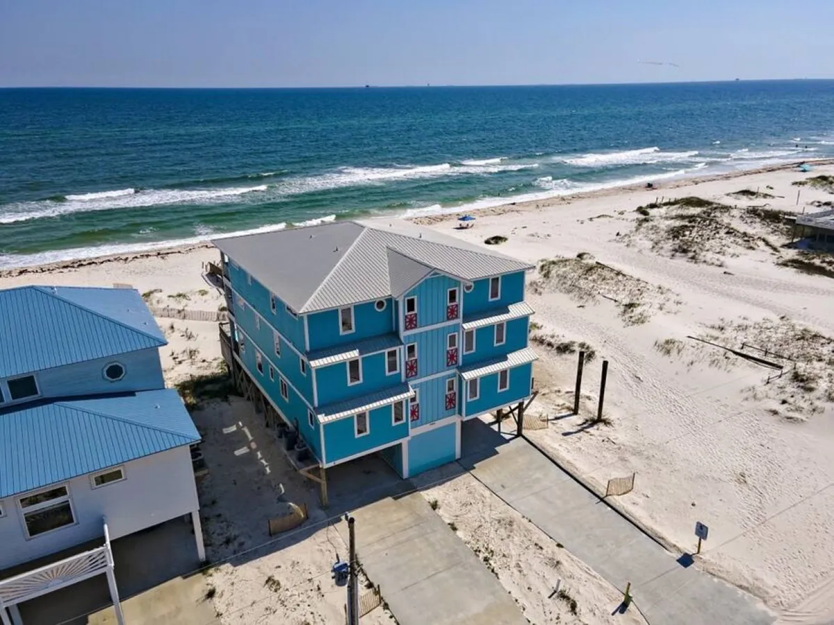 A wide shot of the beachfront property, its turquoise exterior facing the ocean, with gentle waves and a sandy beach in the foreground. The spacious property and open surroundings suggest seclusion and plenty of room for parking or event logistics.