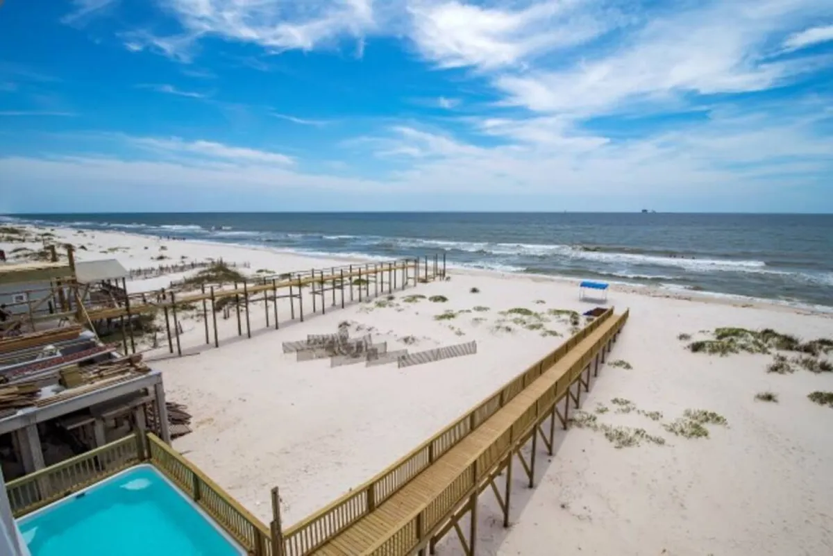This exterior image captures a stunning beachfront view, with a wooden walkway leading to the sand and ocean, as well as a pool area adjacent to the house. The scene is serene, inviting, and showcases the proximity to the beach and the outdoor amenities available for group relaxation.