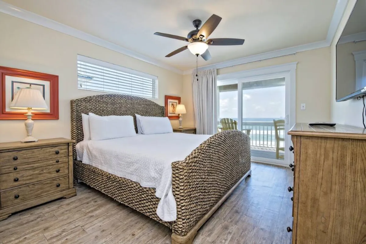 This image showcases a king-sized bed placed near a large window with an ocean view. A nightstand with a lamp and a dresser add functionality, while soft lighting and an inviting bed create a serene, restful atmosphere. The scene exemplifies the tranquil and spacious feel of the upscale bedrooms described in the section, with visual links to private balconies and Gulf views.
