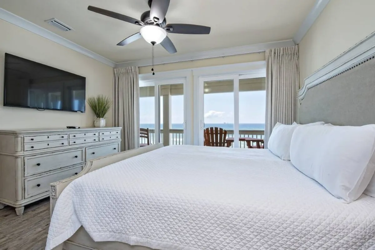 A restful bedroom with a bed dressed in white linens, a large window that frames a sweeping ocean view, a wall-mounted television, and a dresser. There’s a sense of spaciousness and luxury, with the view and balcony access hinting at the exclusivity and relaxation described in the section.