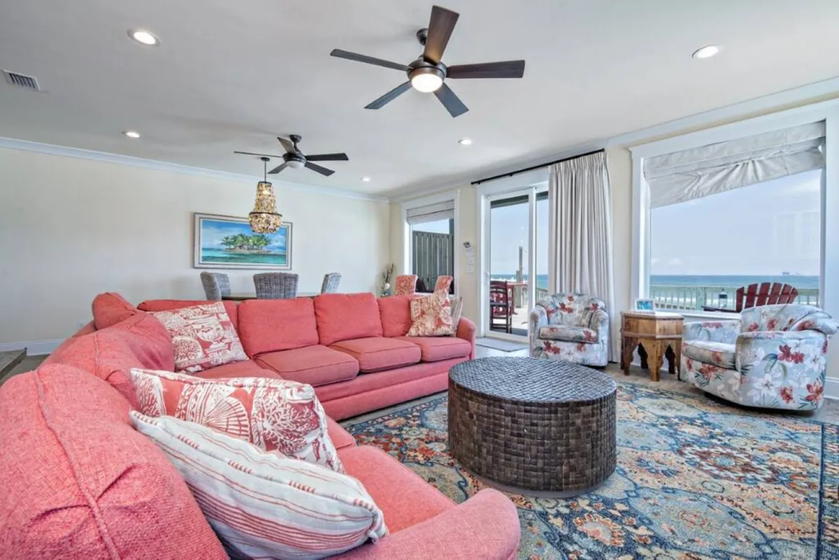 A spacious open-plan living area with a large pink sectional sofa, woven coffee table, and colorful floral armchairs. The room features expansive windows framing panoramic ocean views, with sunlight streaming in to highlight the inviting seating. The setup is ideal for gatherings, with plenty of space for socializing, games, or relaxing while enjoying the Gulf scenery.