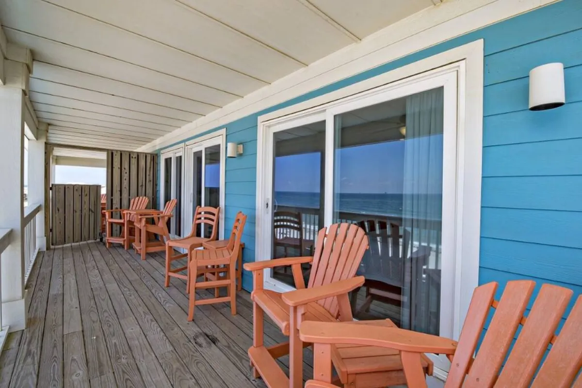 An exterior view featuring adirondack chairs on a spacious deck with blue siding and glass doors, offering an inviting place to relax and socialize while enjoying ocean views and privacy.