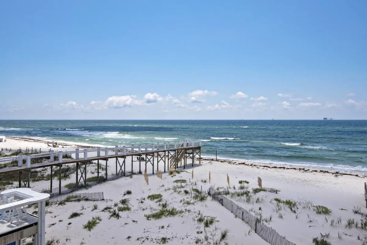 A wooden boardwalk meanders through grassy dunes to a broad beach, with the expansive ocean visible beyond. The dune landscape suggests proximity to natural preserves and wildlife.
