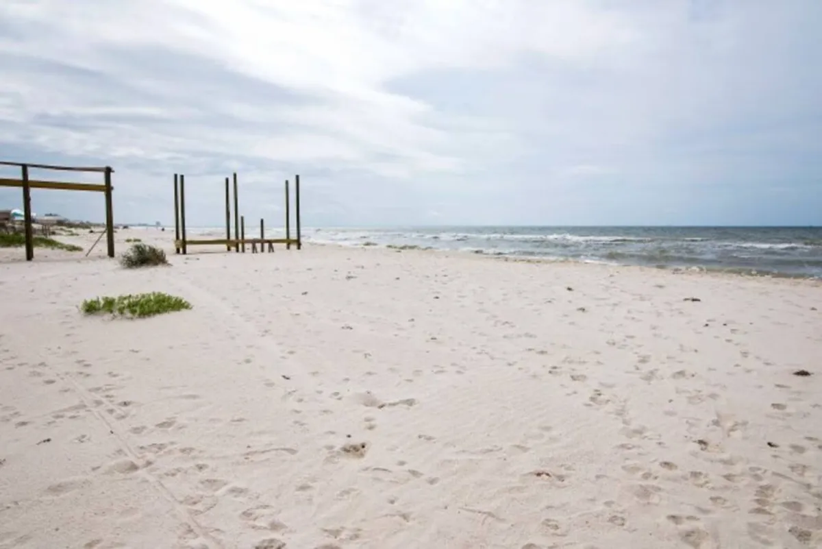 A wide, open view of a sandy beach with gentle ocean waves and simple wooden posts. The unspoiled sand and clear water make it ideal for swimming, sunbathing, and family activities such as building sandcastles.