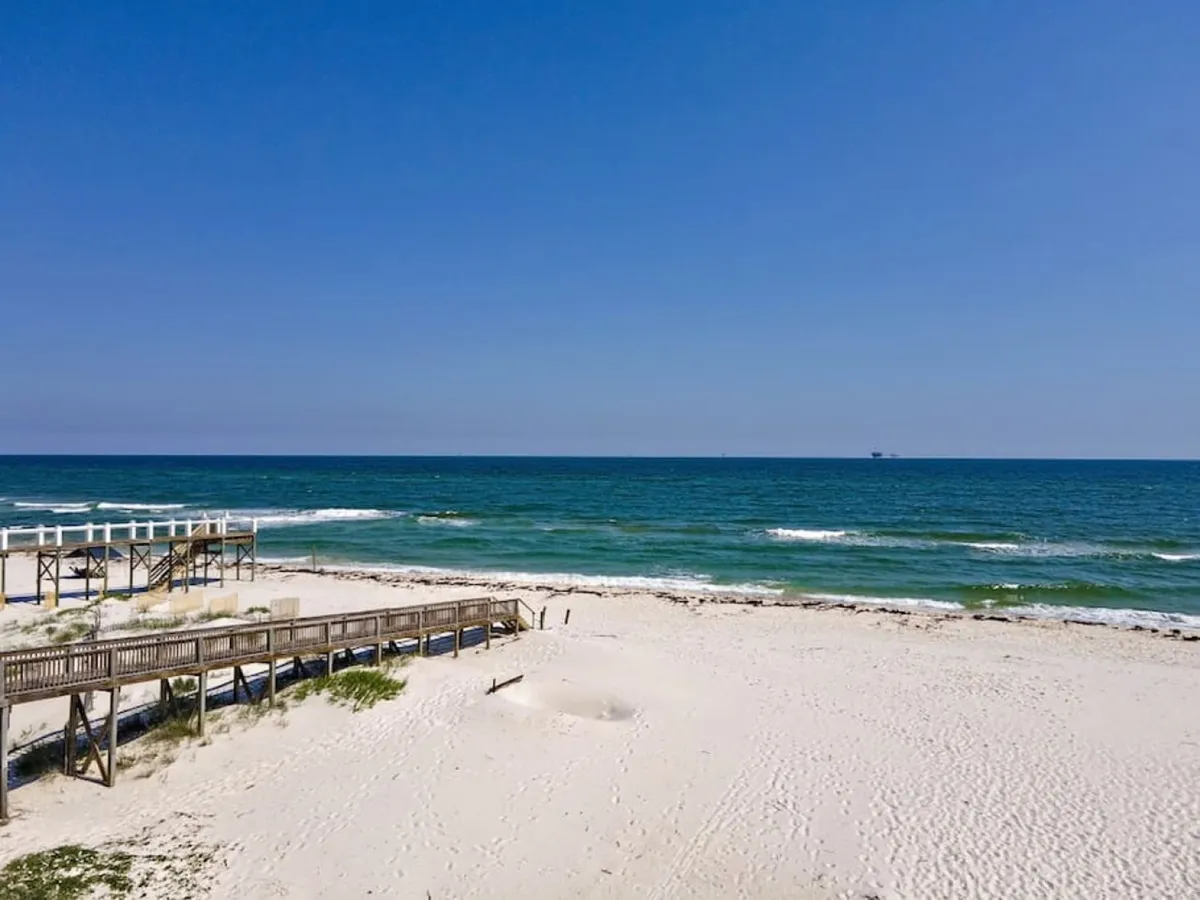 A serene exterior view featuring a wooden boardwalk leading directly to a quiet, sandy beach with gentle ocean waves in the background. The natural dune grasses and open sky set a peaceful, secluded atmosphere.