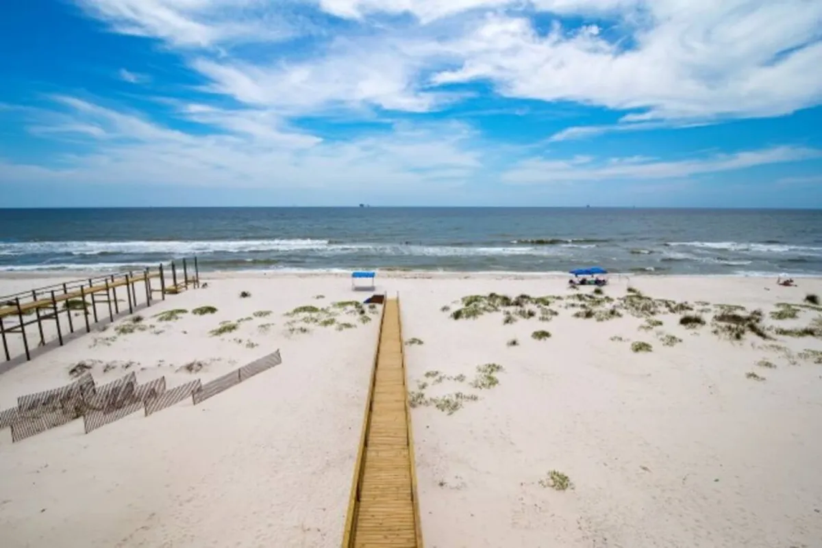 An exterior view of the beach, ocean, boardwalk, and sky. The scene highlights the expansive shoreline and the tranquil blue water, suggesting the dramatic and intimate setting available for events and photos.